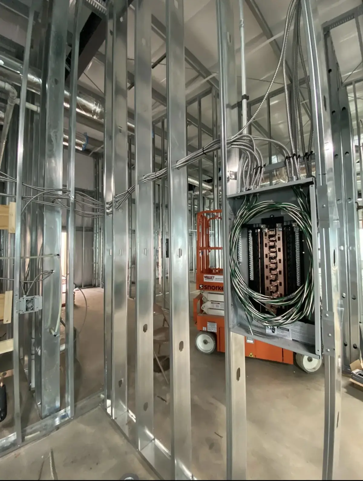 Steel-stud commercial wall with an open panel enclosure during rough-in; cables bundled into the panel, scissor lift in the background