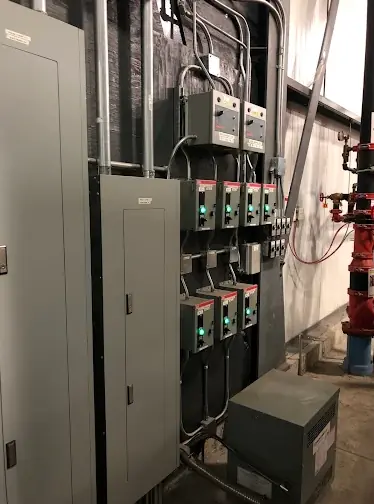 Commercial electrical room with a bank of motor starters and disconnects on a wall, a transformer below and a panel to the left