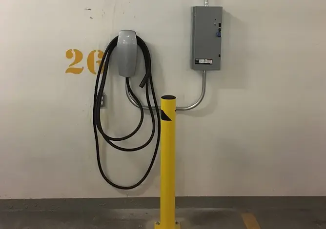 Wall-mounted EV charger with coiled cable in an underground parking spot, with a disconnect box above and a yellow bollard in front